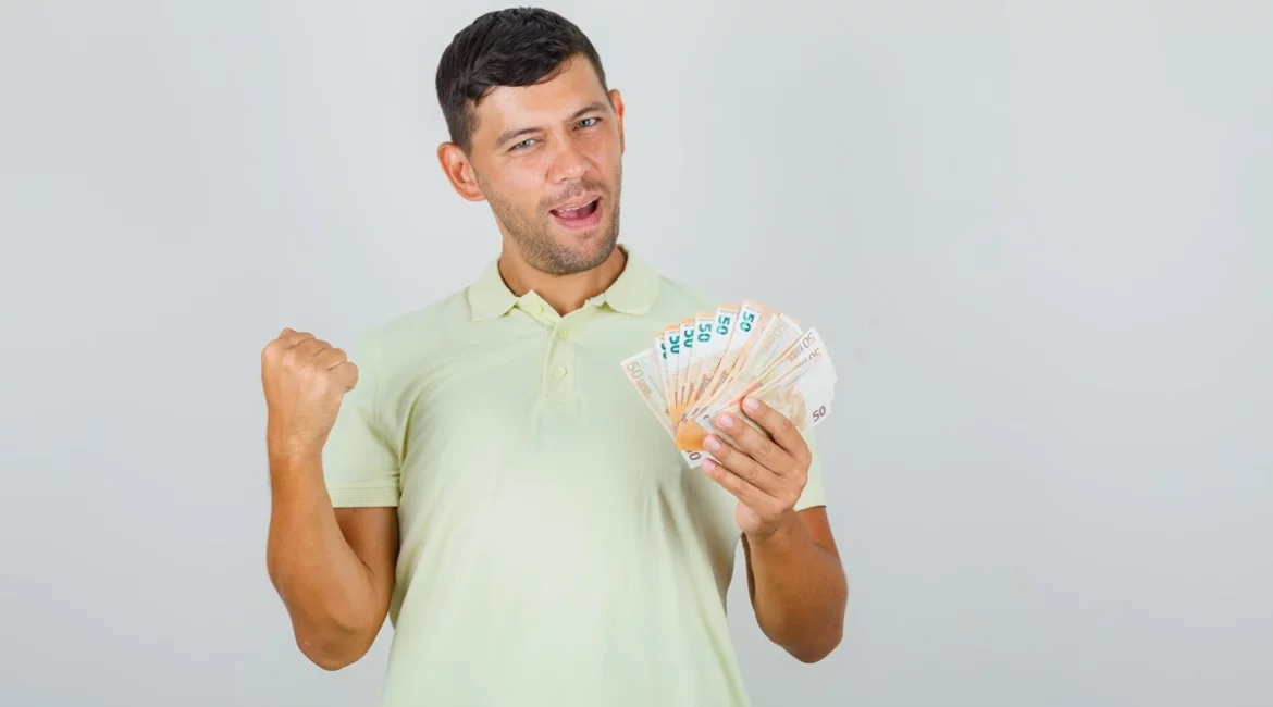 Homem jovem usando camiseta polo clara, segurando várias notas de dinheiro e expressando alegria com gesto de comemoração.