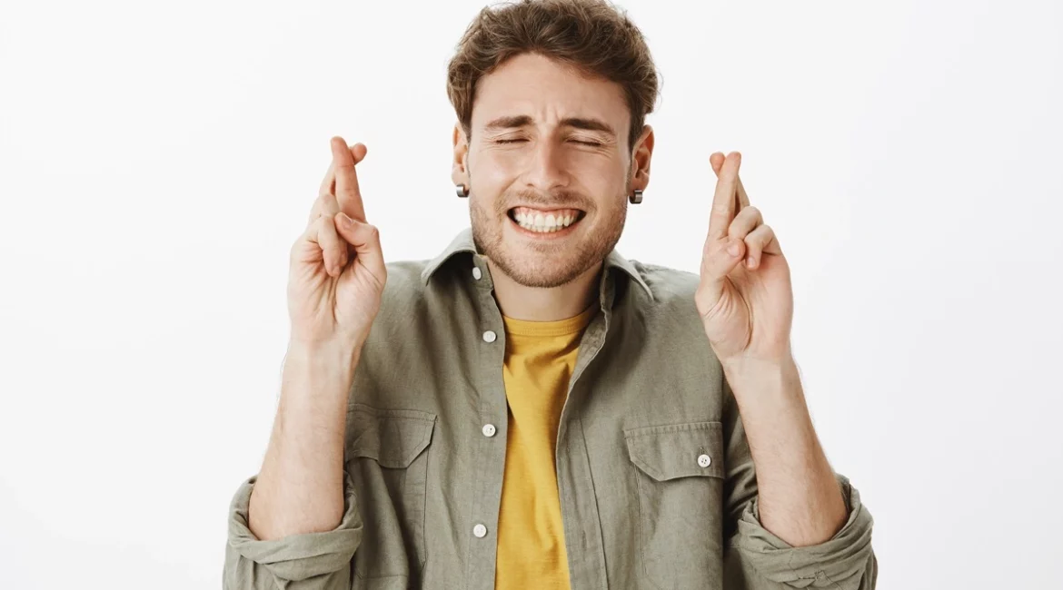 Homem jovem sorrindo com os olhos fechados e dedos cruzados, usando camisa verde e camiseta amarela, expressando confiança e esperança.