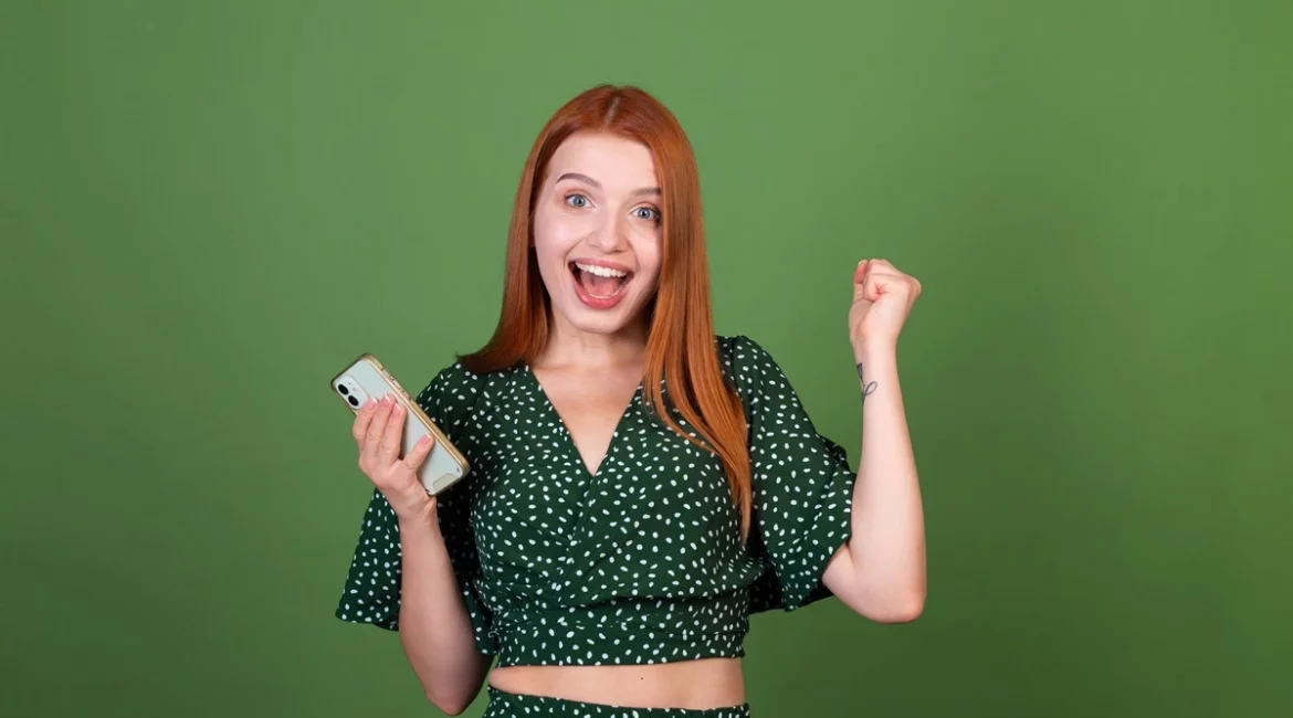 Mulher jovem de cabelo ruivo segurando um celular e comemorando, com expressão de felicidade em fundo verde.