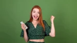 Mulher jovem de cabelo ruivo segurando um celular e comemorando, com expressão de felicidade em fundo verde.