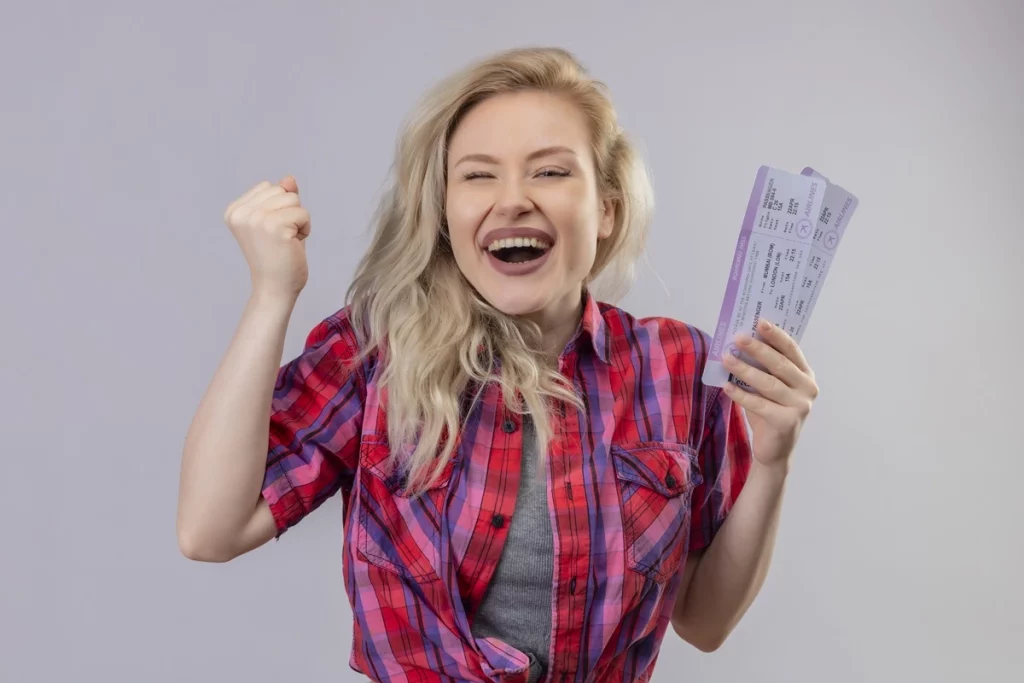 Mulher loira sorridente usando camisa xadrez vermelha, comemorando enquanto segura passagens de viagem.