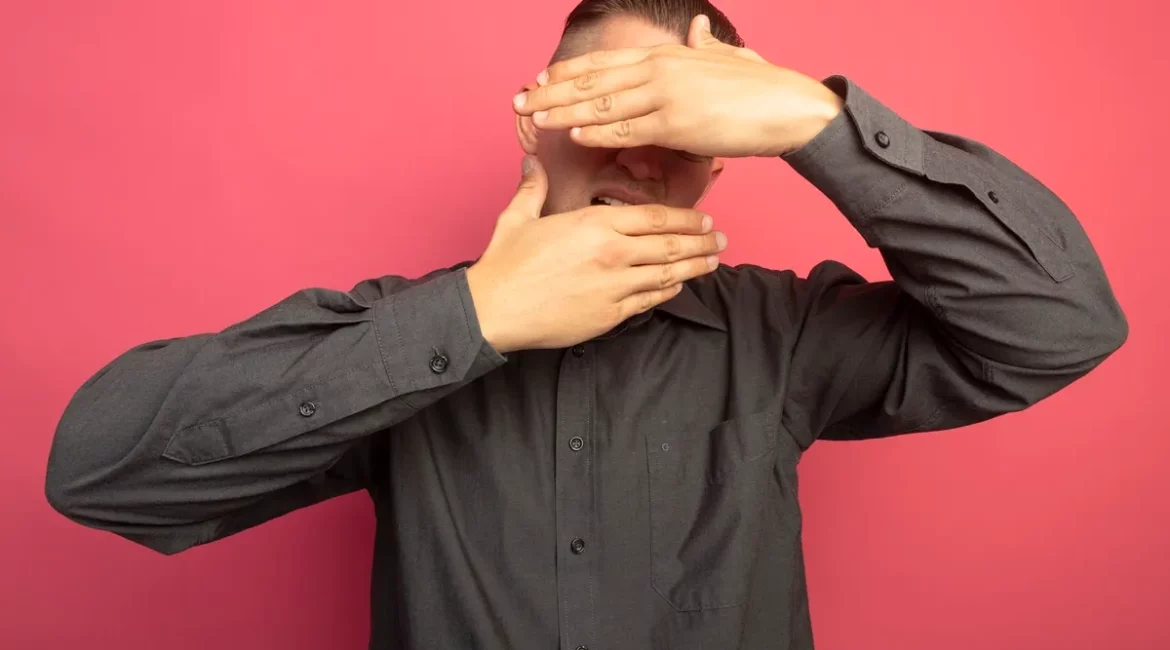 Homem vestindo camisa social cinza escuro, cobrindo parcialmente o rosto com as mãos em um fundo rosa.