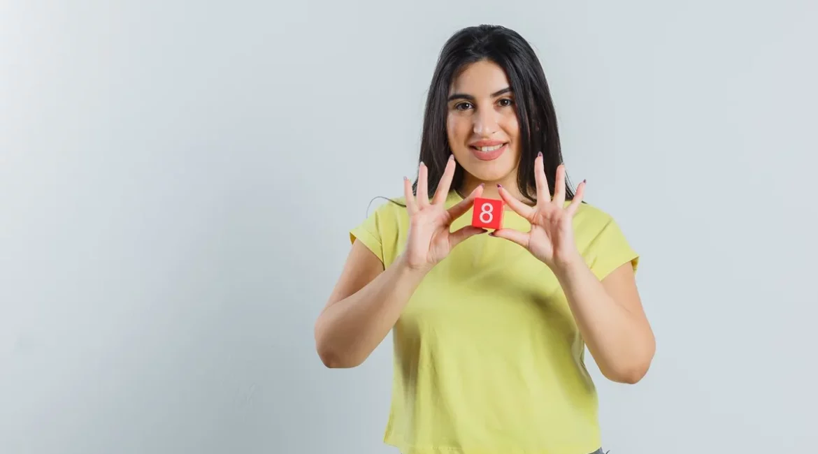 Mulher de blusa amarela segurando um cubo vermelho com o número 8, simbolizando sorte ou escolha de número em jogos e loterias.