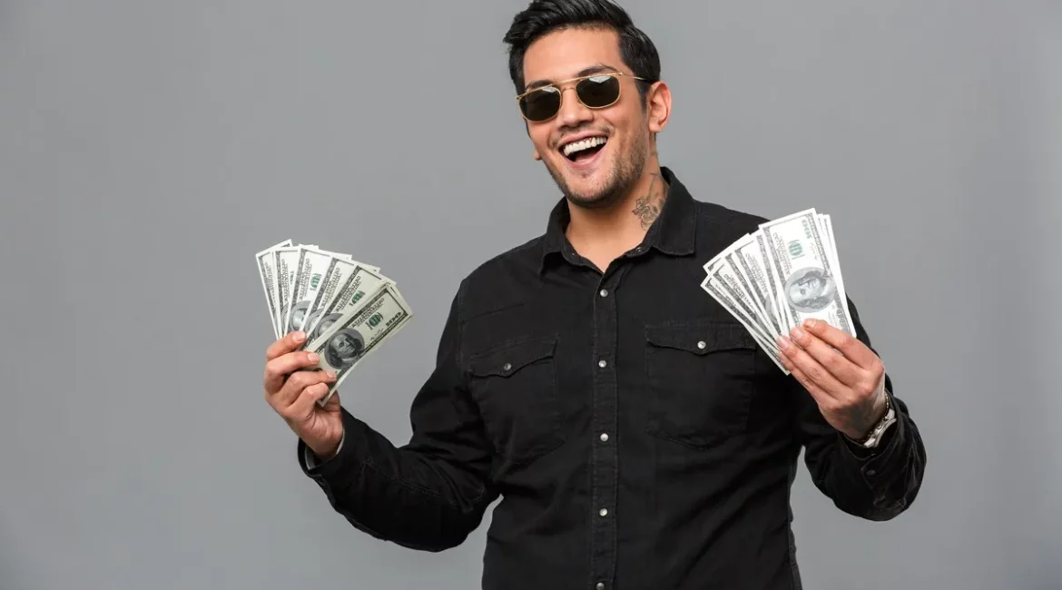 Homem jovem, sorridente, usando óculos de sol e camisa preta, segurando dois leques de notas de 100 dólares, um em cada mão. Fundo cinza uniforme.