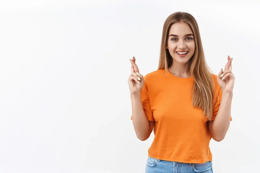 Jovem com blusa laranja e calça jeans, sorrindo e cruzando os dedos em sinal de sorte. Fundo branco minimalista.